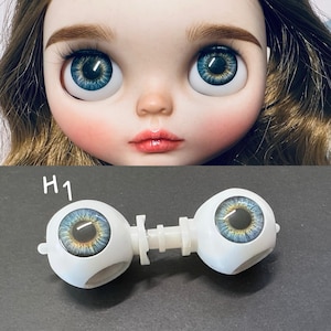 May include: A close-up of a doll's face with large, blue eyes and a fair complexion. Below, a pair of doll eyes with a white connecting piece are displayed. The eyes have a blue iris with yellow and black details. The text "H1" is visible.