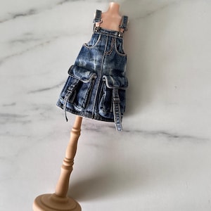 May include: A miniature denim overall dress featuring a distressed dark blue wash, two large front cargo pockets with flaps, and adjustable shoulder straps. The garment is displayed on a small, light-coloured wooden mannequin stand.