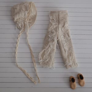 May include: A cream-colored baby outfit set, including a bonnet with ties, pants, and two small shoes. The pants and bonnet have a textured, fringed design. The shoes are a light brown color.