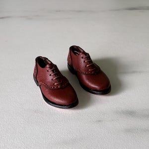 Può includere: Un paio di scarpe oxford in miniatura in pelle marrone con superficie testurizzata, punte arrotondate e sottili lacci marroni. Le scarpe presentano cuciture visibili lungo i pannelli laterali e una suola marrone scuro, pensate come accessori.