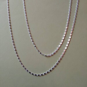 Puede incluir: Dos collares plateados con un diseño delicado y en capas. Los collares presentan pequeños eslabones circulares y planos que captan la luz. Las cadenas se muestran sobre un fondo suave y neutro, destacando su simple elegancia.