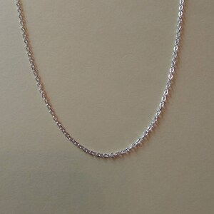 Puede incluir: Un delicado collar de cadena plateada. La cadena está compuesta por pequeños eslabones ovalados interconectados. El collar se muestra sobre un fondo neutro, resaltando su elegante sencillez. Una pieza de joyería clásica.