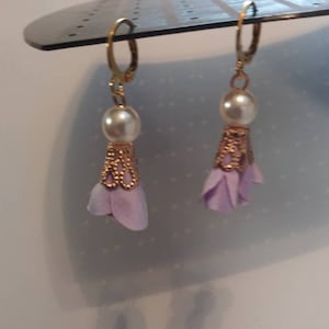 Op de afbeelding: Een paar lavendelkleurige bloemeoorbellen met gouden accenten en een parel aan elke oorbel.