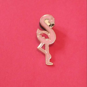 Puede incluir: Pin de esmalte de flamenco rosa con contorno dorado. El flamenco está de pie sobre una pierna con la cabeza inclinada hacia abajo.