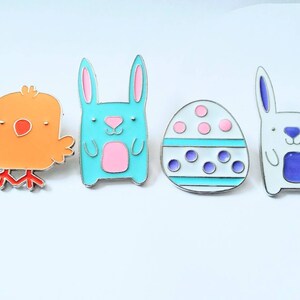 May include: Four enamel pins featuring Easter-themed designs: a blue bunny, a yellow chick, a white egg with pink and purple polka dots, and a white bunny with purple ears and a purple belly.
