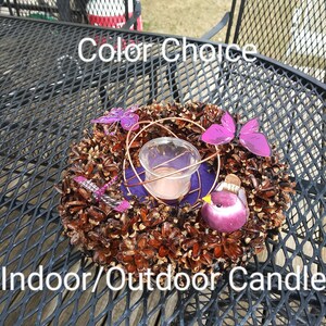 May include: A purple bird and butterfly candle holder made of pine cones and a glass votive. The holder is on a black metal table with the text "Color Choice" and "Indoor/Outdoor Candle" visible.