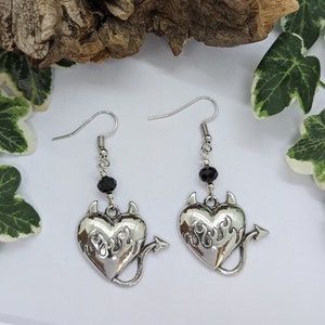 May include: A pair of silver dangle earrings with a black heart charm. The heart has a flame design and devil horns and tail. Each earring has a black bead.