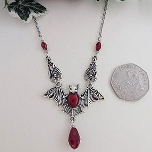May include: A silver-toned bat pendant necklace with red accents. The bat has outstretched wings and a central red stone. The necklace features a chain with red stone details and a teardrop-shaped red stone pendant.