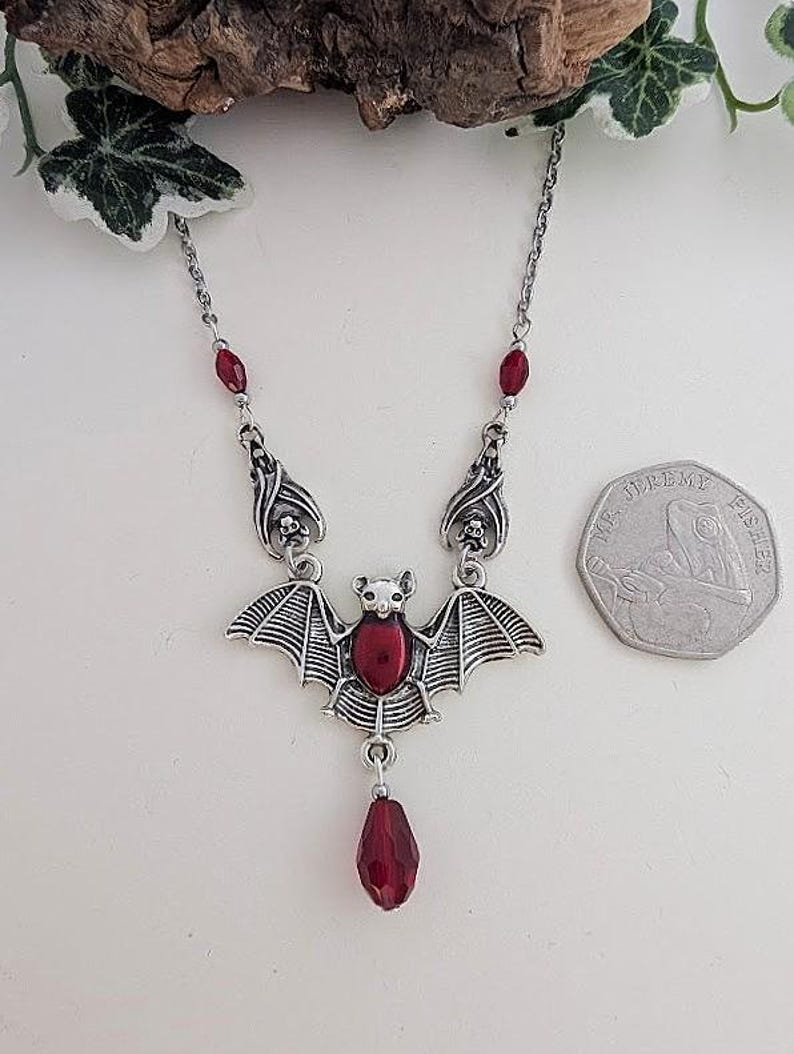 May include: A silver-toned bat pendant necklace with red accents. The bat has outstretched wings and a central red stone. The necklace features a chain with red stone details and a teardrop-shaped red stone pendant.