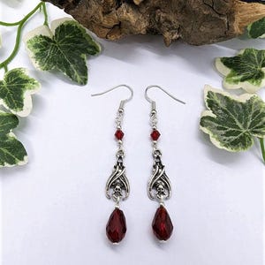 May include: A pair of silver-toned dangle earrings. Each earring features a decorative skull design, red crystal accents, and a large, faceted, teardrop-shaped red crystal. The earrings are displayed on a white surface with decorative greenery.