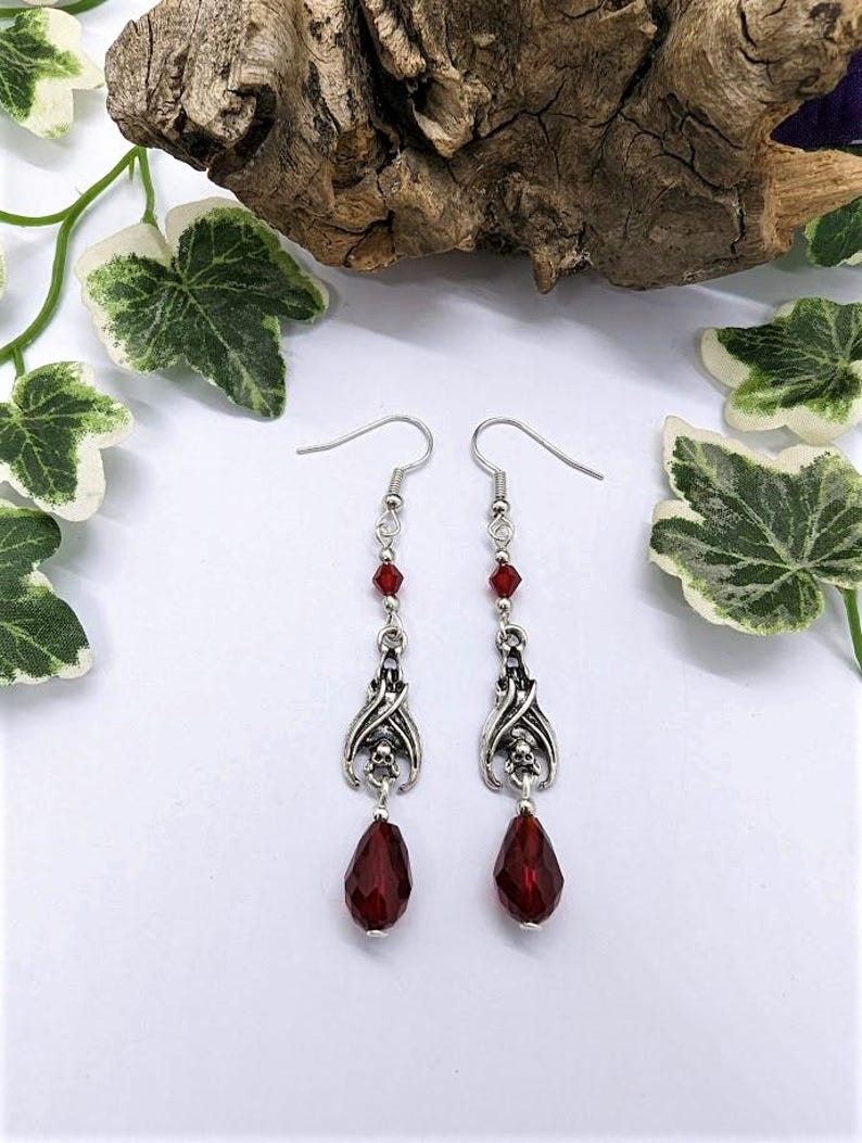 May include: A pair of silver-toned dangle earrings. Each earring features a decorative skull design, red crystal accents, and a large, faceted, teardrop-shaped red crystal. The earrings are displayed on a white surface with decorative greenery.