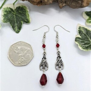 May include: Silver-toned dangle earrings with a gothic theme. Each earring features a bat wing design with a skull, red crystal beads, and a teardrop-shaped red crystal. The earrings are displayed with a coin and greenery.