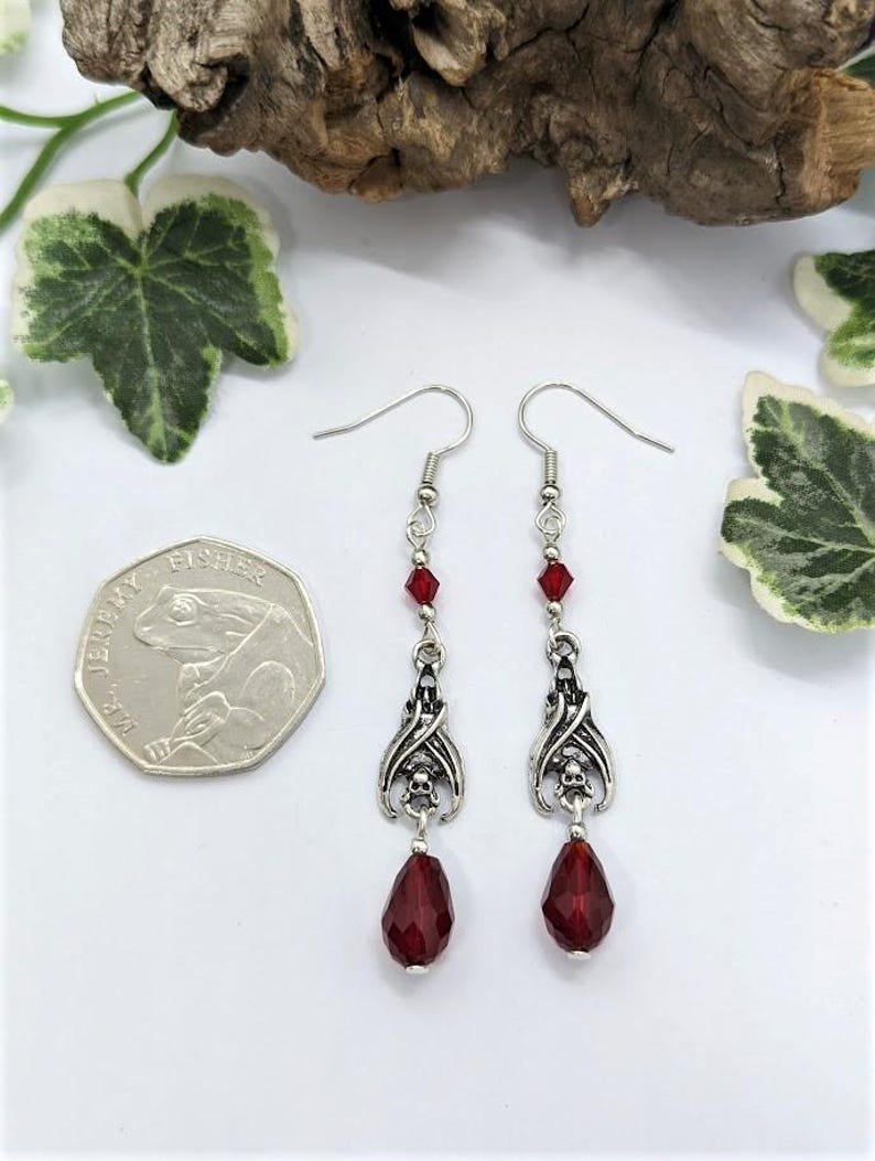 May include: Silver-toned dangle earrings with a gothic theme. Each earring features a bat wing design with a skull, red crystal beads, and a teardrop-shaped red crystal. The earrings are displayed with a coin and greenery.