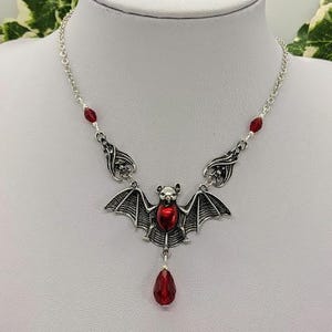 May include: Silver-toned bat pendant necklace with a red center stone and a dangling red teardrop-shaped bead. The necklace has a delicate chain with red bead accents. The bat has detailed wings and a small head.