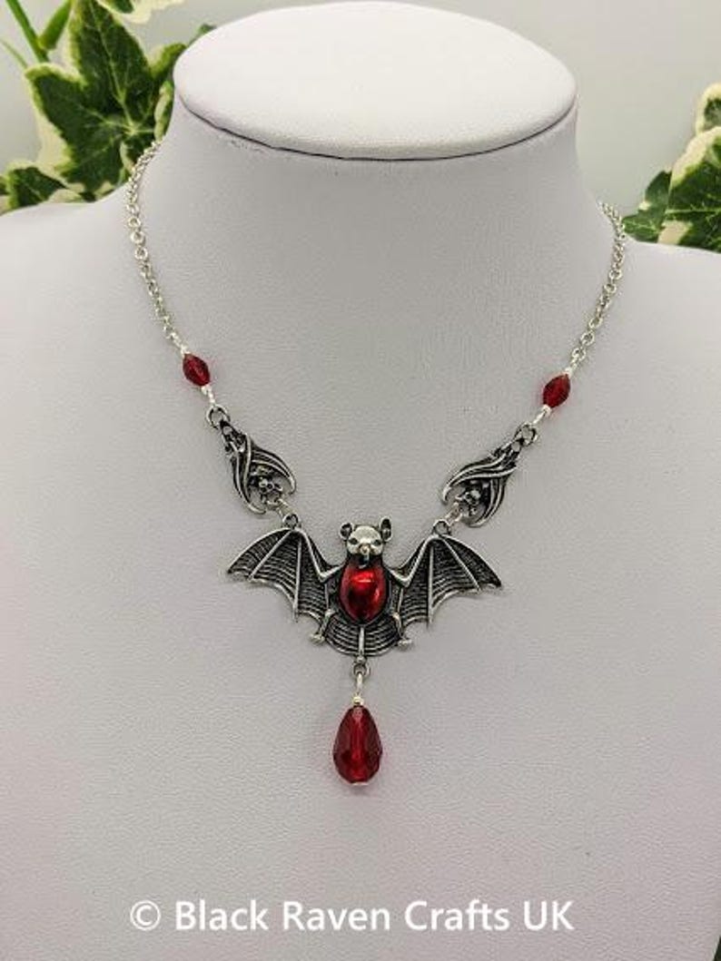 May include: Silver-toned bat pendant necklace with a red center stone and a dangling red teardrop-shaped bead. The necklace has a delicate chain with red bead accents. The bat has detailed wings and a small head.