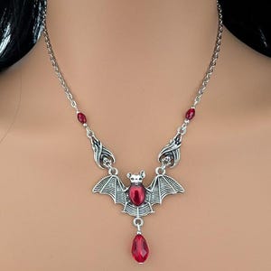 May include: Silver-toned bat pendant necklace with a red gemstone center and a teardrop-shaped red gemstone drop. The necklace has a delicate chain and small red gemstone accents.