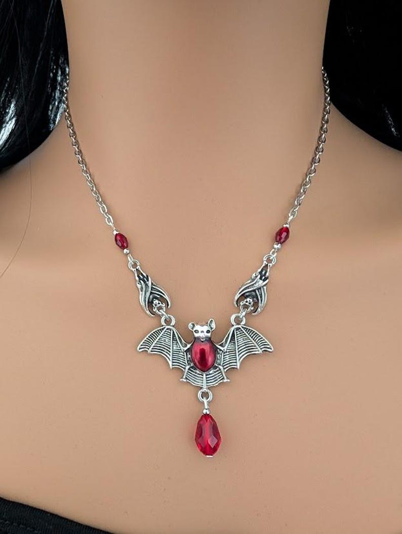 May include: Silver-toned bat pendant necklace with a red gemstone center and a teardrop-shaped red gemstone drop. The necklace has a delicate chain and small red gemstone accents.