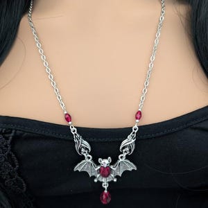 May include: Silver-toned bat pendant necklace with a chain. The bat has outstretched wings and a red crystal in the center. Additional red crystals are set along the chain. The necklace is worn on a black top.