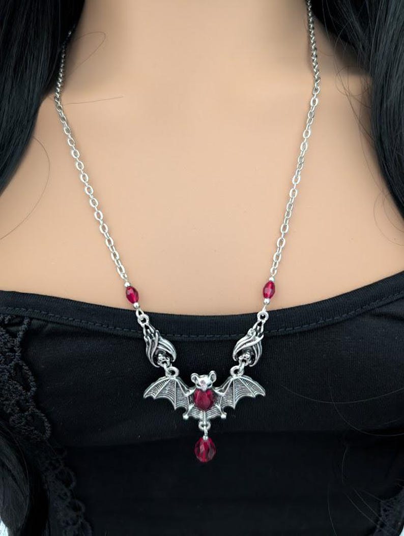 May include: Silver-toned bat pendant necklace with a chain. The bat has outstretched wings and a red crystal in the center. Additional red crystals are set along the chain. The necklace is worn on a black top.