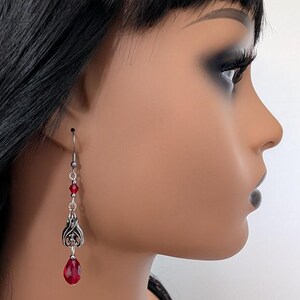 May include: Silver-toned dangle earrings with red teardrop-shaped gems. The earrings feature a decorative design with red beads and a detailed metalwork element. The earrings are shown on a mannequin.
