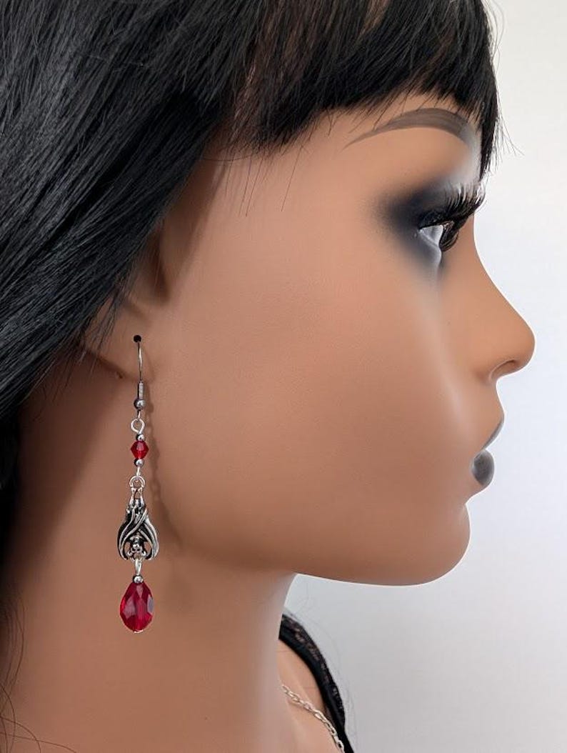 May include: Silver-toned dangle earrings with red teardrop-shaped gems. The earrings feature a decorative design with red beads and a detailed metalwork element. The earrings are shown on a mannequin.