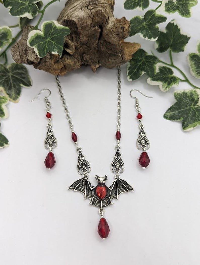 May include: A silver-toned bat pendant necklace with matching earrings. The bat pendant and earrings feature red teardrop-shaped gems and skull accents. The necklace and earrings are displayed on a white surface with green and white foliage.