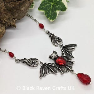 May include: Silver-toned bat pendant necklace with a large red gemstone in the center. The bat has detailed wings and is suspended from a delicate chain with red bead accents. A red teardrop-shaped gem hangs below the bat.