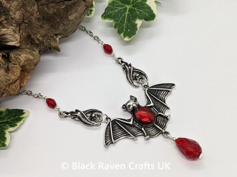 May include: Silver-toned bat pendant necklace with a large red gemstone in the center. The bat has detailed wings and is suspended from a delicate chain with red bead accents. A red teardrop-shaped gem hangs below the bat.