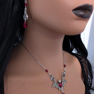 May include: A silver-toned bat pendant necklace and matching earrings with red gemstone accents. The necklace features a bat pendant with outstretched wings and a dangling red teardrop-shaped gem. The earrings have a similar design with red beads.