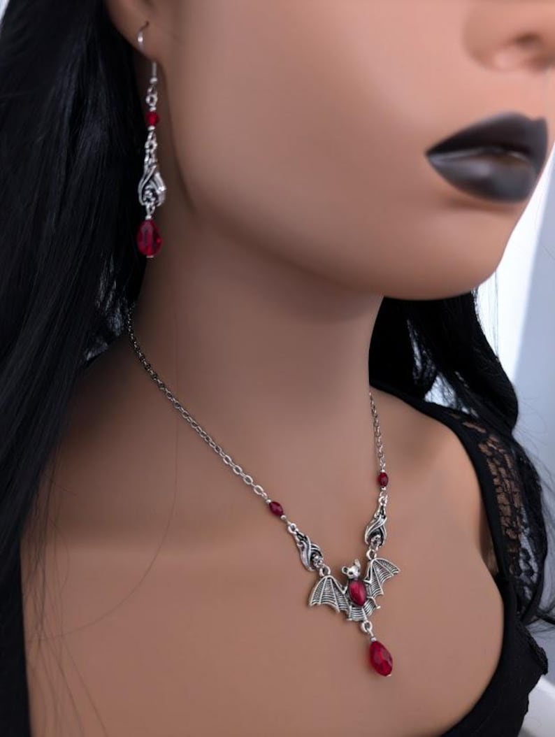 May include: A silver-toned bat pendant necklace and matching earrings with red gemstone accents. The necklace features a bat pendant with outstretched wings and a dangling red teardrop-shaped gem. The earrings have a similar design with red beads.