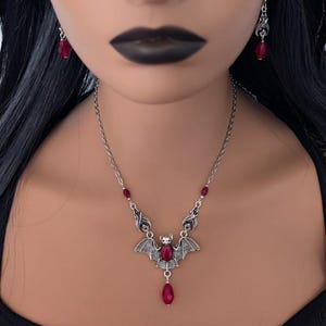 May include: A silver-toned bat pendant necklace with matching earrings. The bat pendant features a large, red, teardrop-shaped crystal and smaller red crystals. The necklace and earrings are worn by a person with black hair and dark lipstick.