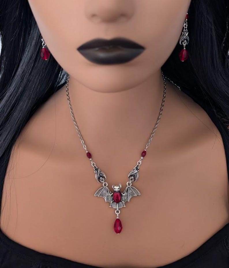 May include: A silver-toned bat pendant necklace with matching earrings. The bat pendant features a large, red, teardrop-shaped crystal and smaller red crystals. The necklace and earrings are worn by a person with black hair and dark lipstick.