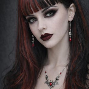 May include: A woman with dark hair, red highlights, and dark makeup. She wears a silver necklace with a bat pendant and red gemstone, matching earrings, and dark lipstick. The overall aesthetic is gothic.
