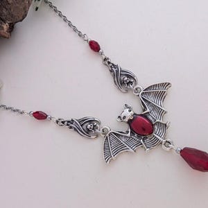 May include: Silver-toned bat pendant necklace with a red gemstone center and a dangling teardrop-shaped gem. The bat's wings are detailed, and the necklace features additional red bead accents.