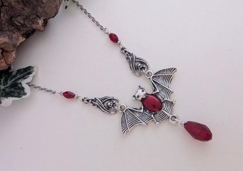May include: Silver-toned bat pendant necklace with a red gemstone center and a dangling teardrop-shaped gem. The bat's wings are detailed, and the necklace features additional red bead accents.