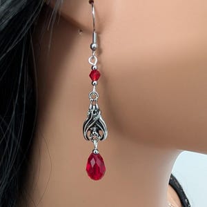 May include: Silver-toned dangle earrings with a gothic bat wing design and a teardrop-shaped red crystal. The earrings also feature small red beads and a silver hook closure. The earrings are shown on a person's ear.