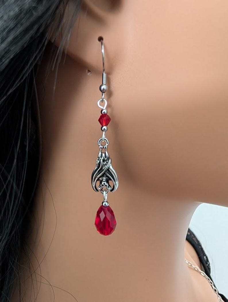 May include: Silver-toned dangle earrings with a gothic bat wing design and a teardrop-shaped red crystal. The earrings also feature small red beads and a silver hook closure. The earrings are shown on a person's ear.