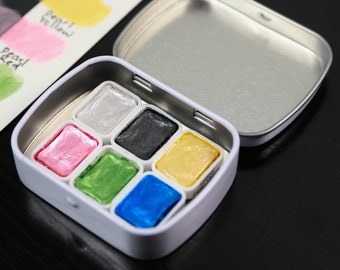 May include: A white metal watercolor paint set with six vibrant colors: pearl, black, pink, green, yellow, and blue. A swatch card displays paint colors with handwritten labels. The set includes a silver metal lid.