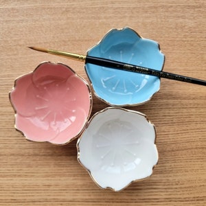Puede incluir: Tres paletas de pintura de cerámica con forma de flor en rosa, azul y blanco, cada una con un borde dorado. Un pincel Winsor & Newton Extra Fine Poster Colour descansa sobre la paleta azul.