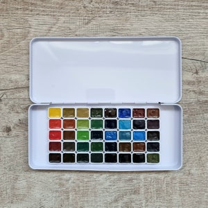 May include: An open white metal watercolor paint set with 36 rectangular paint pans. The colors include yellow, red, brown, green, blue, and brown hues. The set is displayed on a wooden surface.