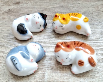 May include: Four ceramic cat figurines in various colors: white with black spots, yellow and orange stripes, blue and white, and brown and white stripes. Each cat is in a sleeping pose with a smiling face. The figurines are on a wooden surface.
