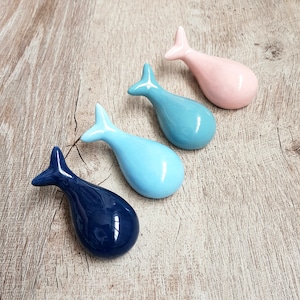 Może przedstawiać: Cztery ceramiczne obiekty w kształcie wielorybów w odcieniach niebieskiego i różowego ułożone są na drewnianej powierzchni. Wieloryby mają błyszczące wykończenie i są zaprojektowane z zakrzywionym korpusem i płetwą ogonową. Kolory obejmują granatowy, jasnoniebieski i różowy.
