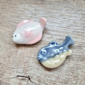 Puede incluir: Dos figuras de peces de cerámica. Uno es rosa y blanco, el otro azul y crema. Ambos son pequeños y parecen ser objetos decorativos, posiblemente para un tema costero o náutico. Probablemente pintados a mano.