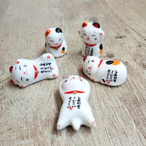 Könnte beinhalten: Fünf weiße Keramik-Maneki-Neko-Figuren mit schwarzen, orangefarbenen und roten Akzenten. Jede Katze hat ein rotes Halsband und japanischen Text. Die Katzen befinden sich in verschiedenen Posen, darunter sitzen, liegen und mit erhobenen Pfoten.