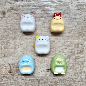 Może przedstawiać: Pięć ceramicznych podpórek do pałeczek z postaciami Sumikko Gurashi. Podpórki są białe, żółte, niebieskie i zielone, ze szczegółami takimi jak czerwona kokarda, żółte uszy i twarz kota. Ułożone są na jasnobrązowej drewnianej powierzchni.