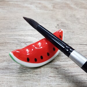 Pode incluir: Um pincel com cabo preto repousa sobre um suporte de cerâmica em forma de fatia de melancia. O suporte é vermelho com detalhes de sementes pretas e casca verde. O pincel tem uma virola prateada e cerdas escuras.
