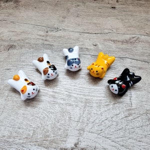 Op de afbeelding: Vijf keramische eetstokjessteunen in katvorm in verschillende kleuren. De katten zijn wit, oranje, geel, zwart en grijs, met gedetailleerde gezichtskenmerken. Elke kat ligt op zijn rug met zijn poten in de lucht.