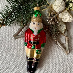 May include: A Christmas ornament shaped like a toy soldier, featuring a red jacket with gold accents, green trim, and black boots. The soldier has a white beard, a green and gold crown, and is suspended by a gold loop. The ornament is set against a backdrop of greenery and decorative elements.