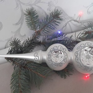 Puede incluir: Un adorno blanco para la parte superior del árbol de Navidad con un diseño de purpurina plateada. El adorno tiene una punta larga y puntiaguda y dos secciones redondas y bulbosas. El diseño incluye purpurina plateada y un patrón decorativo. El fondo incluye una rama de árbol de Navidad y luces.