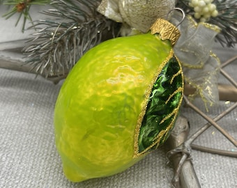 Op de afbeelding: Een kerstversiering in de vorm van een limoen met een getextureerd, heldergroen oppervlak. Een donkergroen bladerontwerp met gouden glitterrand accentueert één kant. De ornament heeft een goudkleurige metalen dop en een zilveren lus.